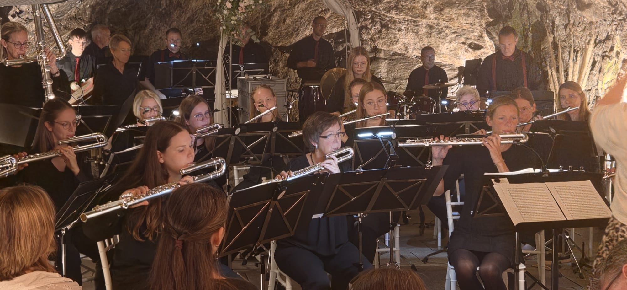 Orchester spielt in der kleinen Höhle. Man sieht erhobene Flöten, eng stehende Schlagwerker und die Wände der Höhle. Alles ist feierlich beleuchtet.